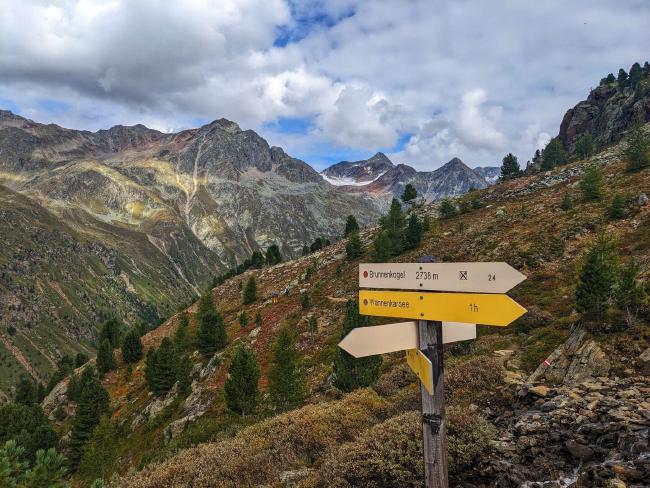 Ein gelbes Wanderschild das zum Brunnenkogel zeigt. Dahinter herbstliche Berghänge mit ein paar wenigen Bäumen. Ganz hinten erkennt man kaum mehr nennenswerte Ausläufer des Stubaier Gletschers