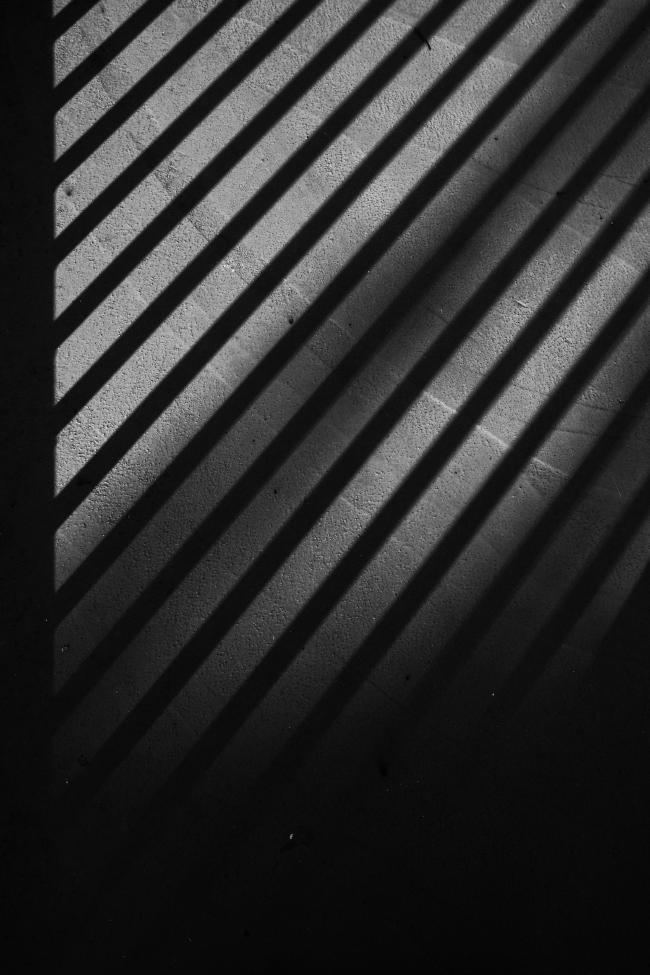 Schwarzweiß Bild im Portrait Format. Zu sehen sind von links nach rechts gehende diagonale Linien die Links in einem breiten Vertikalen Balken starten und rechts aus dem Bild laufen. Der untere Teil (ca. ein Drittel) des Bildes verläuft ins Schwarze. Zwischen den Schatten sieht man den von der Sonne angestrahlten Betonn-Boden der ein Wischmuster aufweist. Das deutlichste Muster ist dabei quasi ein viertel Kreis der das obere Bilddrittel nach rechts hin abtrennt.