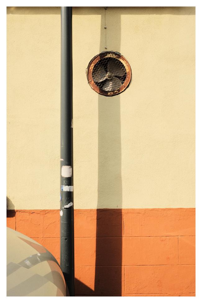 Farbbild im Portrait Format mit weißem Rahmen. Zu sehen ist eine Laternenmast vor einer Hauswand. Diese hat unten Orange gestrichene Steine und oben eine hell-gelbe Fassade. Der Laternenmast wirft darauf einen Schatten. Im oberen Drittel, als wäre es selbst an diesem Schattenmast befestigt ist in der Fassade ein verrostetes Gitter mit einem Ventilator dahinter. Links unten in der Ecke sieht man eine Motorhaube.