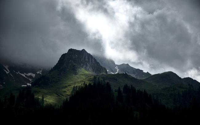 Im Vordergrund sieht man sehr dunkel einige Tannen. Dahinter liegt ein Berggipfel mit einer von der Sonne beleuchteten Gründfläche. Die meisten Teil sind jedoch eher im Schatten. Darüber sind düstere Wolken mit einem Leuchtend hellen Streifen in der Mitte durchden die Grünfläche Beleuchtet wird.