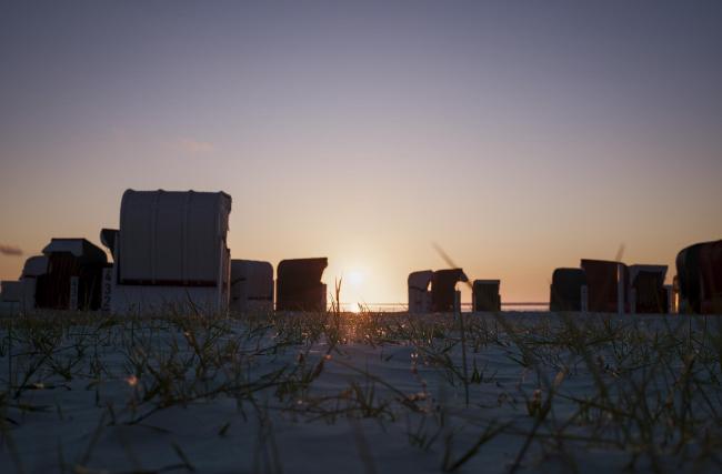 Farbbild im Landscape Format. Das Bild ist aus einer sehr niedrigen Perspektive aufgenommen, wodurch im Vordergrund nicht nur der Sandstrand sondern auch die drauf befindlichen Grashalme zu erkennen sind. Diese glitzern zum Teil orange von der untergehenden Sonne. Dahinter stehen einige verstreute Strandkörbe die mehr als Silhouette als ganz erkennbar sind. Dahinter wiederum liegt ein dünner Streifen Meer, die sehr niedrig stehende Sonne und dann kommt der Himmel in sanften gelb bis blau Tönen.