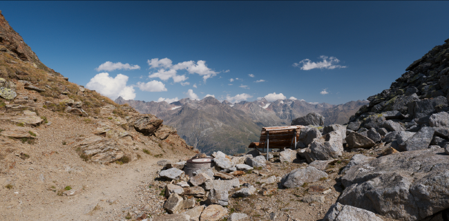 Farbbild in einem etwas breiteren Landscape Format. Der Blich führt über einen Pass und zeigt dahinter die Berge der anderen Tal-Seite. Der Himmel ist Blau mit ein paar Wolken. Im Vordergrund steigen Links und Rechts Berge aus Geröll weiter auf. Links eher gelb und sandig, rechts eher grau und steinig. Leicht rechts der Mitte ist eine große zwei Personen Bank zum liegen auf der man das Panorama genießen könnte.