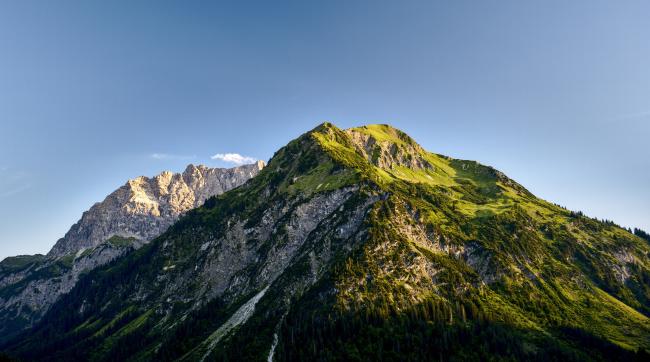 Ein Berg mit vielen grünen Flächen im Vordergrund. Diese werden zum Teil von Bäumen und zum Teil von rohem Fels unterbrochen. Im Hintergrund ist ein Berg nur aus Fels zu erkennen. Der Himmel ist wolkenlos und alles wird seitlich von der untergehenden Sonne angestrahlt.