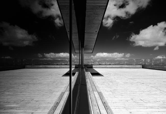 Schwarz-Weiß Bild. Landscape Format. Man sieht eine weitestgehend symmetrische Spiegelung einer holz Bank und Terrasse in einer abgedunkelten Scheibe. Ein Teil im Hinteren Bild liegt im Schatten. Mit Mittelgrund ist die Terrasse durch eine Beschriftete Glaswand begrenzt. Der Himmel wirkt dunkel und die Wolken sehr plastisch. Das Bild ist in den Bergen an einer Bergstation einer Seilbahn aufgenommen.
