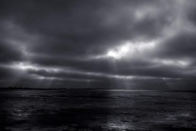 Schwarz-Weiß Bild. Blick über das Watt bei Ebbe. Es ist stark bewölkt aber es scheinen einzelne sichtbare Lichtstreifen durch die Wolken auf das Watt wo sie sich spiegeln. Hier sind es eher viele kleine Lichter.
