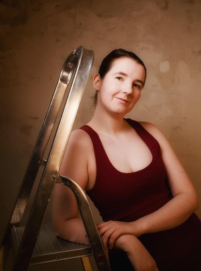 Farbbild im Portrait Format. Zu sehen ist eine Frau mit zusammengebundenen Haaren und einem roten Kleid die mit dem rechten Arm auf einer Leiter lehnt. Das Bild ist etwa auf der Hüfte abgeschnitten. Der Hintergrund ist eine unverputzte ungleichmäßige in der Sonne beige Betonwand. 