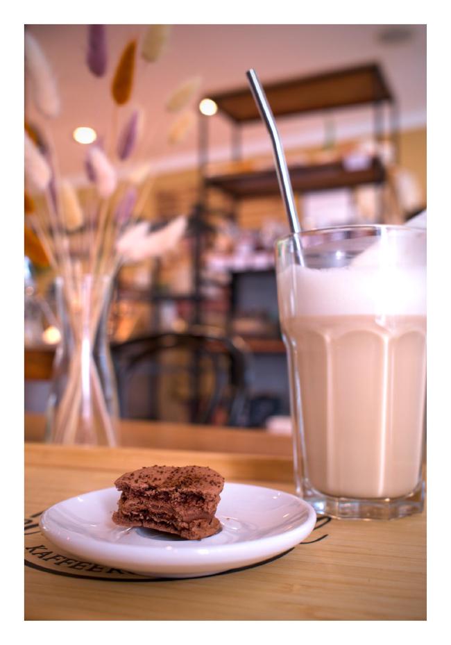 Blick auf ein Tablett mit einem kleinen Teller und einem angebissenen braunen, veganen Caffee Macaron darauf. leicht rechts dahinter ein Pokal Glas mit einem Stahl Strohalm in dem ein Chai Latte mit Hafermilch ist. In der dahinter liegenden Unschärfe sieht man eine Vase mit bunten Gräsern und dahinter die Einrichtung des Cafés 