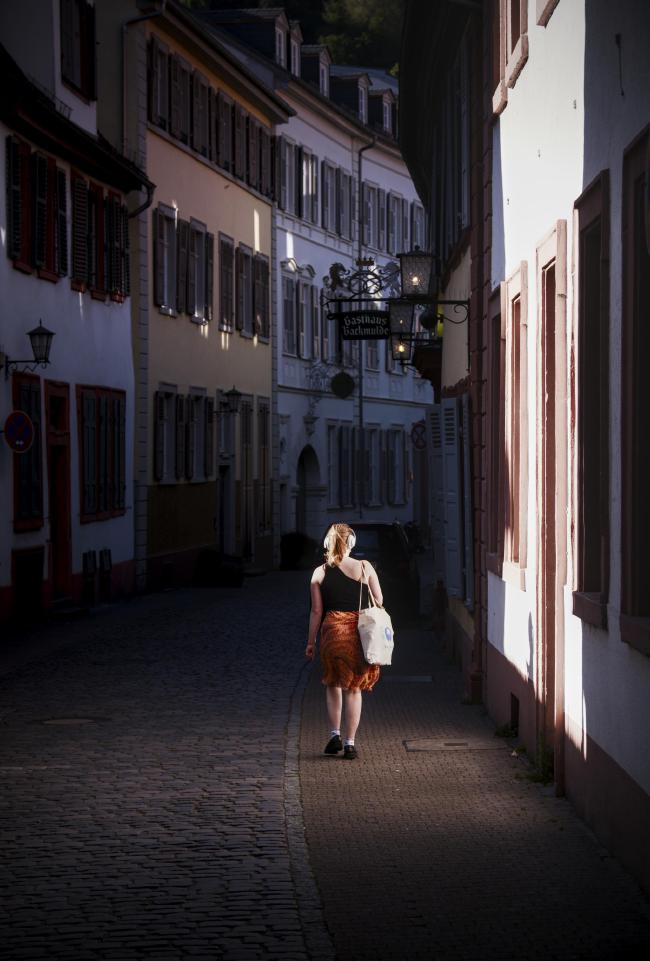Farbbild im Portrait Format. Blick durch eine Gasse in der Altstadt von Heidelberg die schon im Schatten des Abends liegt und daher eher dunkel ist. Aber von Links scheint die Sonne durch eine Lücke auf eine Hauswand und erzeugt durch reflexion einen feinen Lichtschein auf dem Boden. Durch diesen Lichtschein und durch das Licht der Sonne läuft eine Person mit roten Haaren, Kopfhörern, einem schwarzen Oberteil, einer hellen Tragetasche, einem knallig orangenen gemusterten Rock und schwarzen Schuhen. Die Person läuft von der Kamera weg. 