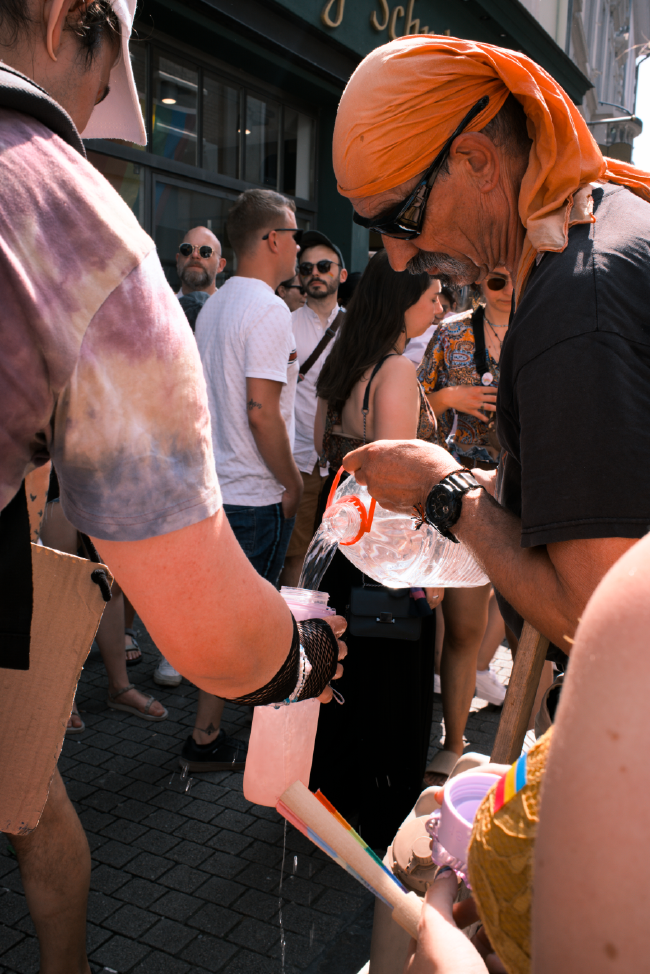 Ein (vermutlich) Mann mit einem orangenen Tuch um den Kopf und Sonnenbrille füllt in der Menge einer Person eine hingehaltene Flasche mit Wasser aus einem größeren Container auf.