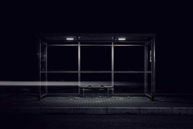 Schwarzweiß-Bild mit leicht bläulichem Einschlag im Landscape Format. Zu sehen ist ein Wartehäuschen von einer Bushaltestelle das nur durch zwei kleine Lampen im Dach beleuchtet wird. Es ist leer. Auf dem gepflasterten Boden liegen ein paar Blätter. Man sieht die Bordsteinkante und davor ein Stück Straße. Der Hintergrund ist nahezu vollkommen Schwarz. Auf Höhe der drei Sitze ist eine Leuchtspur zu sehen die etwa ein drittel von rechts anfängt einzufaden und sich dann über die Sitze bis zum linken Rand zieht. Alles ist ziemlich symmetrisch und mittig zentriert.