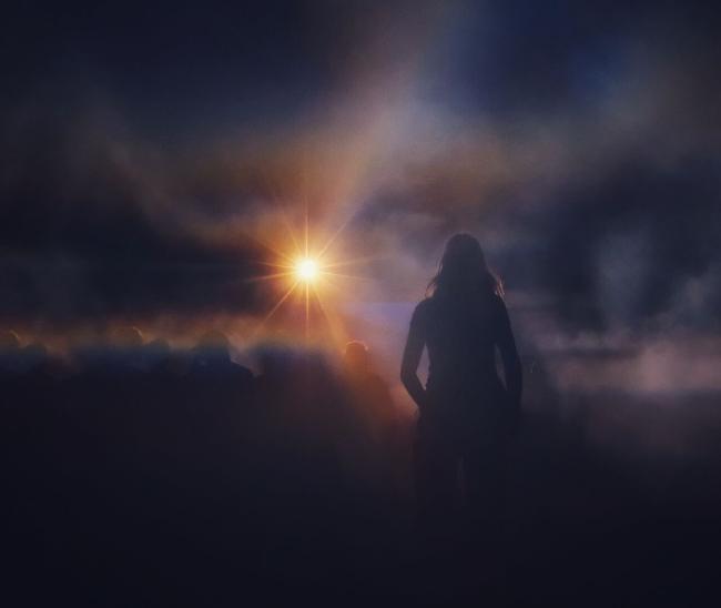 Eine Person mit langen Haaren steht leicht rechts vor einem sehr hellen sternförmigen Licht das durch Nebel leuchtet und regenbogenartige Farbspiele zeigt. Weiter vorne ist noch eine Gruppe zu erkennen. Alle Personen sind nur als Silhouette erkennbar