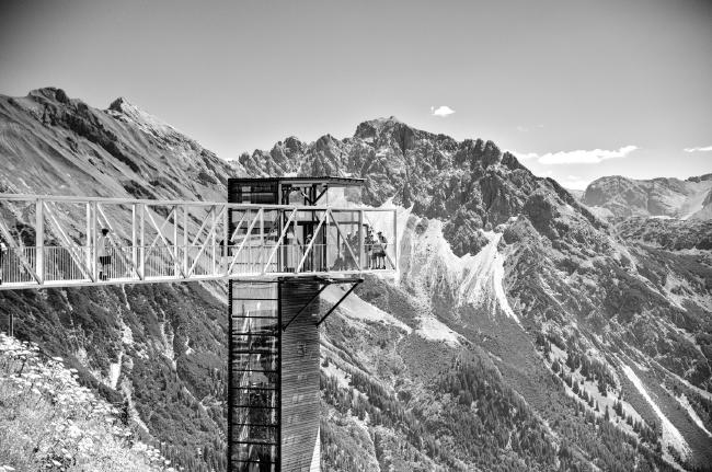 Schwarz-Weiß Bild. Im Hintergrund sind schroffe Berggipfel zu erkennen die weiter unten bewaldet sind. Im Vordergrund sieht man von links bis etwas zur Bildmitte einen farblich eher hellen Steg auf dem einige Menschen stehen. An dessen Ende schließt sich eine weitere Struktur aus Holz und Glas an die eher dunkel in ihren Farben ist und einen Fahrstuhl enthält.