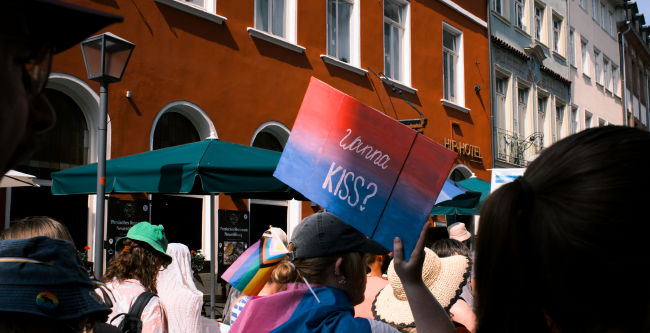 Bild von einer Person in der Heidelberger Innenstadt mit alten Gebäuden die ein Schild in Bi Pride Farben hoch hält auf dem steht: Wanna Kiss?
