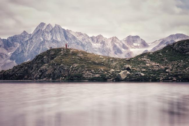 Blick über die Wasserfläche eines Bergsees die durch eine Langzeitbelichtung milchig wirkt. Hinter der Wasserfläche sind sanft geschwungene Hügel des Berges zu sehen auf dem wir und befinden. Am rand links ist es eher felsig und rechts davon mehr leicht grün von der spärlichen Vegetation. Überall liegen unterschiedlich große Felsbrocken. Auf einem der Hügel etwa 1/3 von links steht eine Person mit roter Jacke. Hinter den Hügeln sieht man die kargen Berggipfel der anderen Talseite. Links ist der Rest eines Gletschers zu sehen. Fer Himmel ist bedeckt mit wenig strukturierten Wolken.