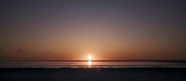 Farbbild in einem Panorama Format. Es zeigt den Sonnenuntergang mit der Sonne gerade noch über dem Horizont, wobei sich die Sonne in der Wasseroberfläche spiegelt. Der Himmel ist fast Wolkenlos und zeigt einen schönen Blau über Gelb zu Rot Farbverlauf. Im Vordergrund ist der Strand als dunkle unregelmäßige Fläche zu sehen. 
