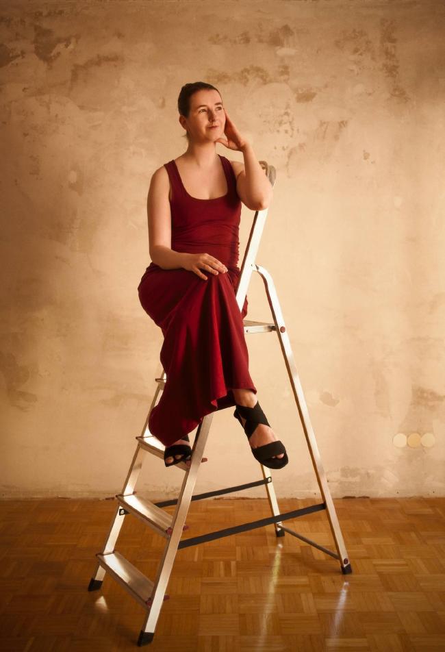 Farbbild im Portrait Format. Zu sehen ist eine Frau mit zusammengebundenen Haaren und einem roten Kleid auf einer Leiter. Die linke Hand ist an ihrem Gesicht und die Beine überkreuzt. Die offenen Schuhe sind schwarz. Der Boden ist ein Mosaik aus Holz und der Hintergrund ist eine von der Sonne angeleuchtete Wand ohne Tapete mit einem interessanten unsauberen Muster das in verschiedenen beige Tönen leuchtet.
