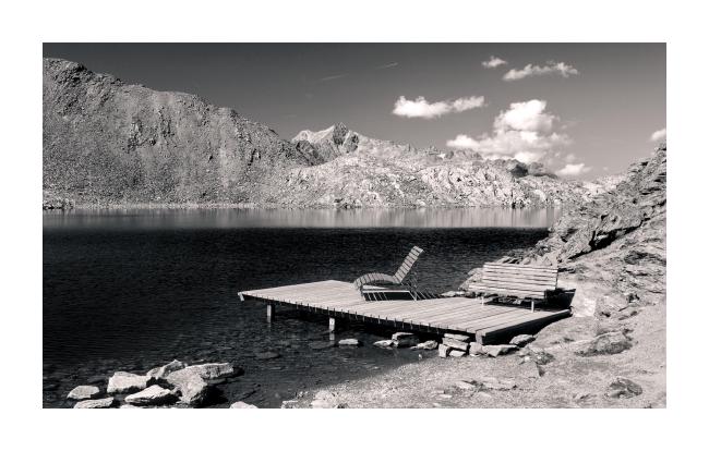 Monochromes Bild mit weißem Rahmen im Landscape Format. Darauf zu sehen ist eine Platform die in einen sehr dunklen fast schwarz wirkenden, aber sehr klaren See hinein ragt und auf der zwei Bänke stehen,. Der See ist umgeben von sehr schroffen Felsen und im Stück Himmel das man sieht sind ein paar kleine Wolken zu erkennen. Das Bild ist nicht exakt schwarz weiß sondern hat einen leicht wärmeren Ton. Durch die starken Kontraste, das Fehlen von Menschen und das dunkle Wasser entsteht eine leicht surreale Stimmung im Bild.