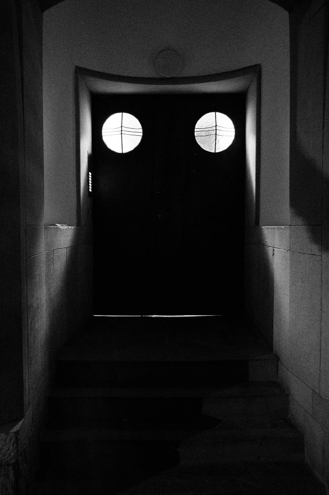 Schwarzweißbild im Hochkant Format. Fotografiert wurde ein Gebäudeeingang mit einer gewölbten Form am Ende. Die dortige Tür hat zwei Flügel und in jedem ein größeres rundes Fenster mit milchigem Glas. Der Flur dahinter ist beleuchtet wodurch Licht durch diese Fenster in den ansonsten dunklen Hauseingang fällt. Die Fenster sind vertikal mit einem und horizontal mit drei Streben in der Mitte verziert. Unter der Tür scheint leicht Licht durch, was zusammen den Eindruck von Augen und Mund erzeugen kann.