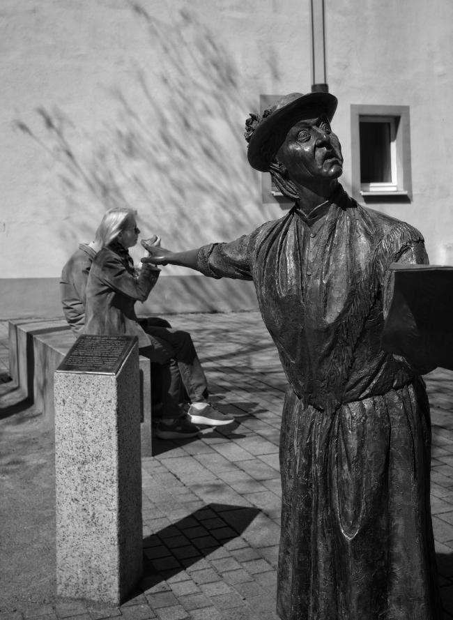 Schwarz-Weiß Bild im Portrait Format. Eine Bronzefigur, die eine ältere Frau mit Hut und langem Kleid zeigt schaut empört und zeigt mit ausgestrecktem Arm und Zeigefinger hinter sich. Dort ist eine Steinbank auf der zwei Personen sitzen. Die sichtbare Frau isst gerade ein Eis und es wird direkt auf den Löffel gezeigt, den sie gerade zum Mund führt. Dahinter ist eine Haus Fassade, mit einem Schatten von einem fast noch blattlosen Baum und zwei Fenstern. Der Boden ist mit rechteckigen Steinen gepflastert.