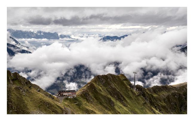 Farbbild mit weißem Rahmen im Querformat. Ein weiterer Blick über die Landschaft und das Montafon. Zu sehen ist im Vordergrund ein Berggrad auf dem in einer Senke die Wormser Hütte liegt. Der Hang ist grün gelb bewachsen. Dahinter liegt zum großen Teil von Wolken verdeckt das Montafon Tal bei Schruns und im Hintergrund sieht man schon die Berge in der Schweiz.