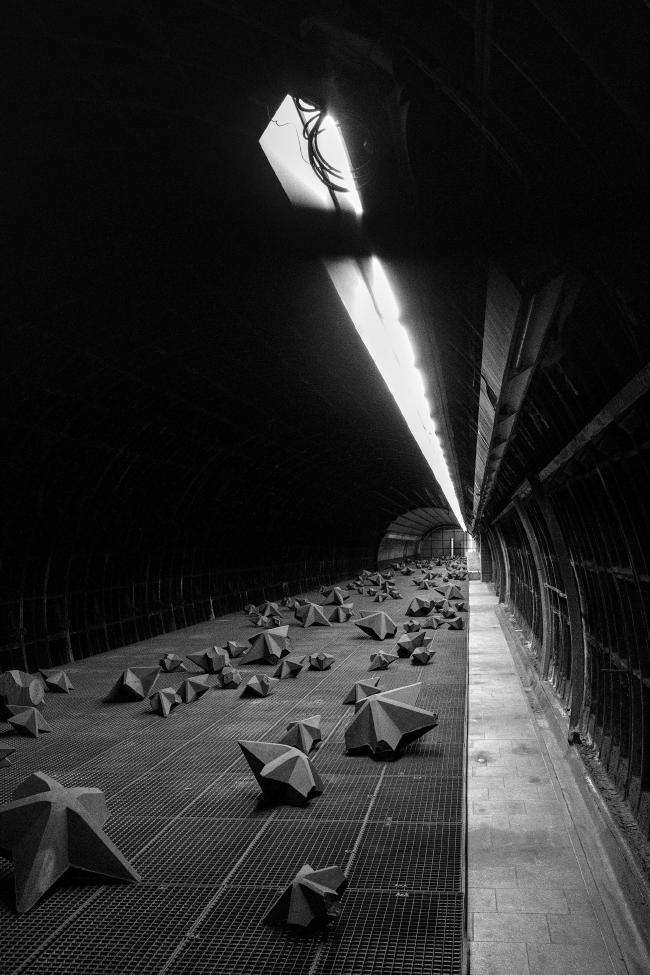 Schwarz-Weiß Bild im Portrait Format. Zu sehen ist eine U-Bahn Röhre die nicht in Betrieb ist. Auf einem Gitterboden liegen dort viele große und kleine zerbrochene Sterne aus Beton. Beleuchtet wird das Ganze von einer Leiste aus Licht die durchgehend im oberen Teil des Bildes zu sehen ist. Der rest der Szene ist sehr dunkel bis schwarz.
