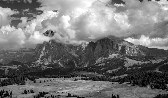 Scharz-Weiß Foto im Landscape Format. Es zeigt den Blick im Sommer über Teile der Seiseralm, eine Fläche aus Landwirtschaftlichen Flächen, kleinen Hütten und Bäumen. Dominiert wird das Bild aber vom Wolkenverhangenen Langkofelmassiv.