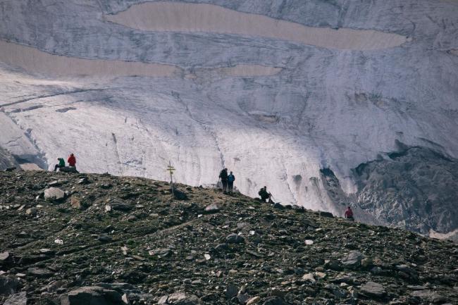 Einige Menschen tummeln sich auf einem Grat aus Geröll und rujen sich vom Wandern aus. Dahinter ragt ein Gletscher empor und bedeckt die ganze obere Hälfte des Bildes. Das Bild ist durch die Aufnahme mit dem Teleobjektiv extrem komprimiert.