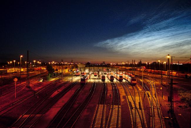 Bild eines Rangierbanhofs mit vielen Laternen. In der Mitte ist eine Wartungshalle und einige Triebwägen zu erkennen die mit einem anderen Licht beleuchtet sind. Der Himme ist dunkel, aber rechts oben sind Nachtleuchtende Wolken zu erkennen.