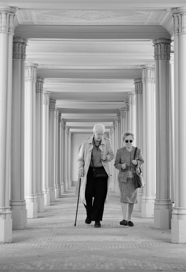 Schwarz-Weiß Bild im Portrait Format. Zwei älter Frauen, eine mit einem Gehstock laufen durch einen Säulengang und unterhalten sich.