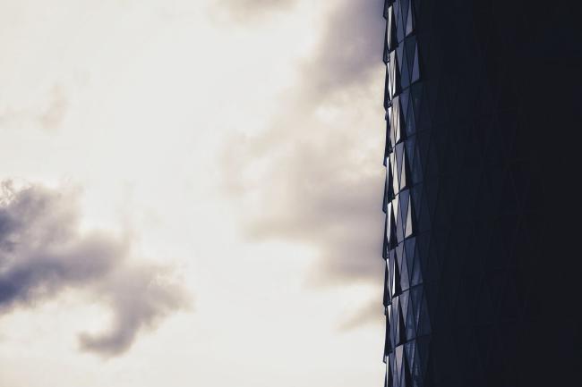 Bild in Landscape Orientierung von einer Hochhausfront aus dreieckigen Scheiben. Das Hochhaus verschwindet rechts im Schatten während die anderen 2/3 bewölkten Himmel zeigen. Einige davon stehen wie Schuppen leicht geöffnet hervor. Bild relativ monochrom allerdings gehen die Farben leicht ind gelb/blau.