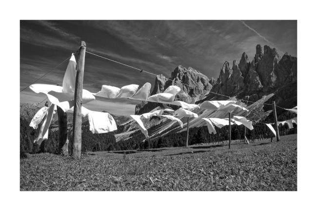 Schwarzweiß-Bild mit breitem weißen Rahmen. Abgebildet ist strahlend weiße Wäsche die auf Leinen die über Holzpfosten gespannt sind hängt und im Wind auf den Betrachter zu weht. Jedoch leicht rechts vorbei. Das ganze steht auf einer Wiese. Dahinter ragen schroffe Berggipfel in den nur leicht bewölkten Himmel.