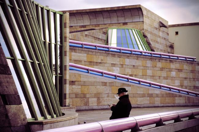 Ein älterer Mann mit Hut steht rauchend an eine rosa Röhre gelehnt. Diese verläuft zusammen mit einer blauen Röhre an verschiedenen Stellen und in verschiedenen Winkeln durchs Bild. Der Blick ist dabei auf die Staatsgalerie Stuttgart gerichtet und man sieht an verschiedenen Stellen grün eingeräumte Fensterfronten 