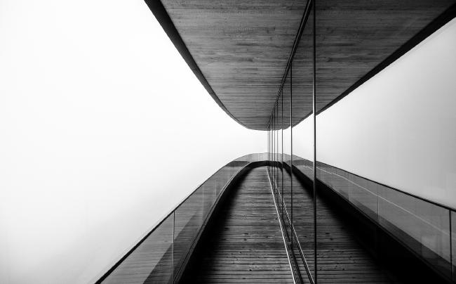 Schwarz-Weiß Bild. Es herrscht dichter Nebel auf dem Bild, der alles entferntere verschwinden lässt. Man sieht eine leere schmale Terrasse mit einem Geländer aus Glas und einem Holzboden. Diese Spiegeln sich genau wie die Decke in einer bodentiefen Fensterfront, sodass eine gewisse Symmetrie gegeben ist.