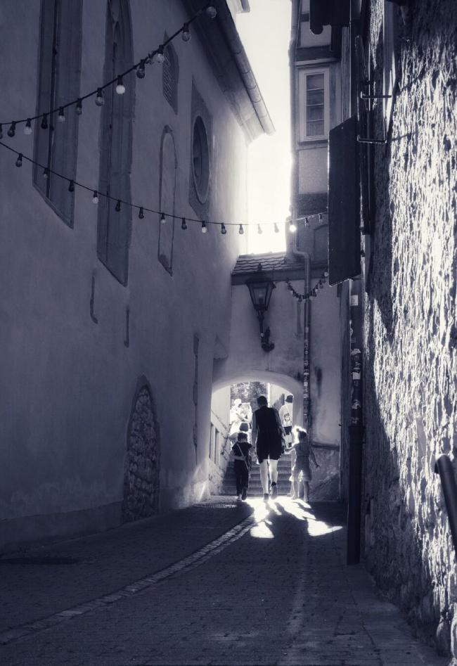 Eine Person mit zwei Kindern geht durch einen Torbogen in einer mittelalterlichen Gasse. Das Licht fällt schräg ein, sodass die Rechte Steinwand beleuchtet ist und auch Licht durch das Tor fällt. Dahinter befindet sich kaum sichtbar eine Treppe und weitere Personen. Die Gasse ist mit einer Lichterkette geschmückt. Das Bild ist hochkant und schwarzweiß mit einer leichten Blau Einfärbung.