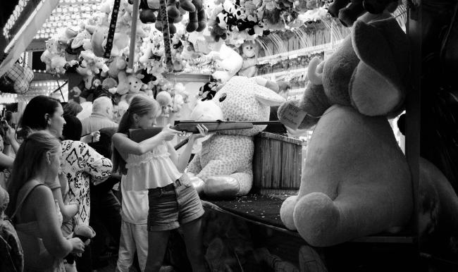 Schwarzweißbild im Landscape Format. Ein Mädchen mit kurzer Hose und weißem Oberteil steht sehr konzentriert mit einem Luftdruckgewehr in einer Schießbude. Es macht den Eindruck, dass sie nicht das erste mal so ein Gewähr hält. Dabei ist sie umgeben von großen und kleinen Kuscheltieren. Hinter ihr stehen weiter Personen die ihr zuschauen. Evl. ihre Familie.