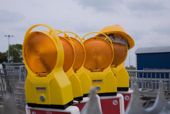 Farbbild im Landscape Format. Ein Reihe von Warnbaken mit gelb-orangenen Warnlichtern darauf die etwas unordentlich in einer Reihe stehen. Von den Warnbaken ist allerdings nur ein kleines oberes Stück zu sehen. Das letzte der 5 Warnlichter hat einen Gelben Baustellenhelm auf und scheint in Richtung der Kamera zu gucken. Im Hintergrund sieht man einen Bauwagen und verschiedene Zaunelemente. Im Vordergrund leicht unscharf im unteren Bildteil ein paar Zaun Spitzen.