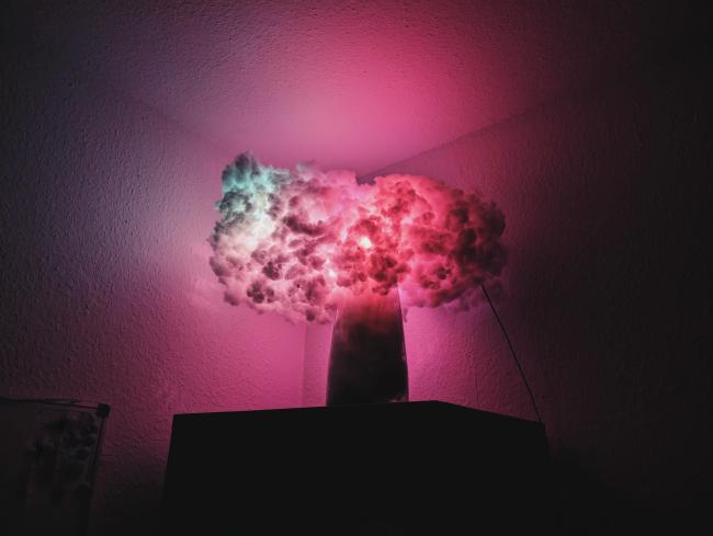 In der Ecke oben auf einem Schrank steht eine Vase auf der eine rot und grün leuchtende Wolke liegt. 