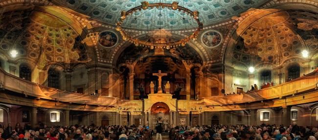 Panorama des Krichenraums der in Gold und Grünen Visuals erstrahlt. Am unteren Rand sieht man das Publikum.