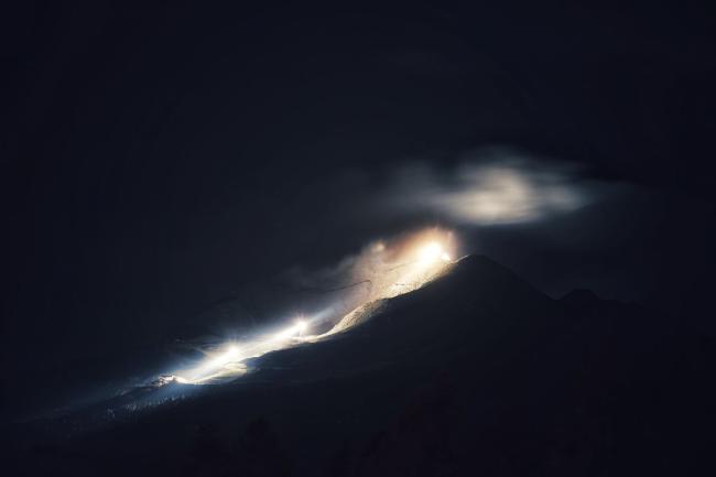 Nachtaufnahme. Der größte Teil des Bildes liegt im Dunkeln. Leicht rechts von fer Mitte ist ein sehr helles, warmweißes Licht, dass einen Berggipfel sichtbar macht. Das Licht wird auch von einer Wolke darüber reflektiert. Weiter unten in einer Art halb offenem Kessel sind nochmals zwei helle Lichter die in der leicht diesigen Luft einen scheint nach links unten haben. Dort befindet sich ein Gebäude, das in den Lichtkegel der hier einige Bäume beleuchtet einen Schatten reißt. Das ganze wirkt ein bisschen wie ein ausbrechender Vulkan. Es ist allerdings eine Seilbahnstation mit Flutlichtstrahlern am Gipfel und den Masten der Bahn. Das Gebäude links unten ist die Mittelstation.