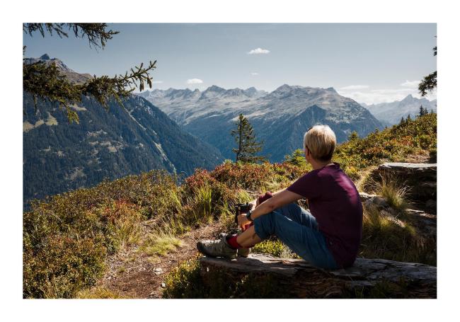 Farbbild im Landscape Format mit weißem Rahmen. Eine Frau mit kurzen blonden Haaren, einer blauen drei-viertel Hose und einem burgunderroten Shirt sitzt auf einer Bank aus groben Baumstämmen und guckt in die Ferne. Dabei hält sie eine Flasche. Um geben ist das ganze von Gras und Blaubeersträuchen. Dahinter sind viele Gipfel auf der anderen Tal-Seite zu sehen. 