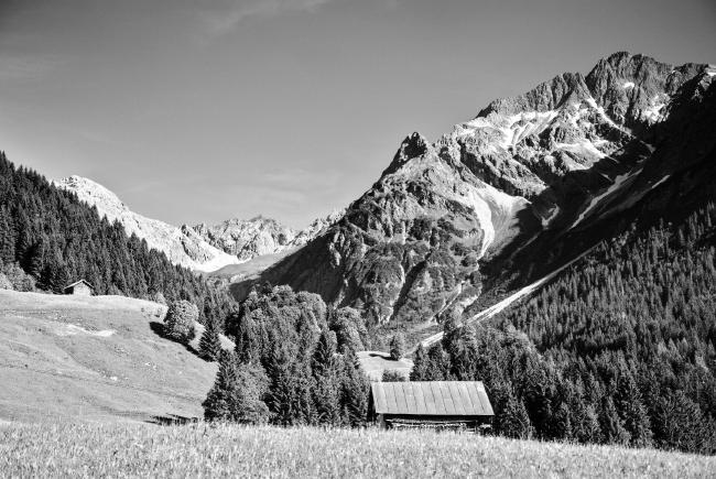 Schwarz-Weiß Bild einer Alpenlandschaft. Im Vordergrund lugt das Dach einer Strohhütte hinter einem Hügel hervor. Darum verteilt stehen einige Bäume und etwas weitere entfernt eine weiter kleine Hütte. Ansonsten sieht man einiges an Wiese. An den Hängen in Mittlerer Entfernung sieht man Bäume stehen, weiter hinten im Bild werden die Berge höher, schroffer, und haben keinen Baumbewuchs mehr. Vereinzelt sieht man noch Schneefelder hin höheren lagen. Am Himmel sind nur einige sehr feine Wolken zu erkennen.