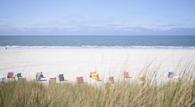Farbbild im Landscape Format. Blick über eine Düne auf einen Strand und das Meer. Das Untere Bildviertel wird von Gras auf einer Düne gefüllt das auch den Abhang zum Strand verdeckt. Direkt am Strandanfang stehen verschiedene bunte aber fast immer leere Strandkörbe. Dahinter liegt viel leerer Strand nur ganz links an der Wasserkante laufen zwei Personen. Dahinter dann das Meer mit sehr kleinen Wellen und der Himmel. Der Strand  macht etwa 1/3 des Bildes aus, das Meer 1/4 und der Himmel dann nochmal etwa 1/3.