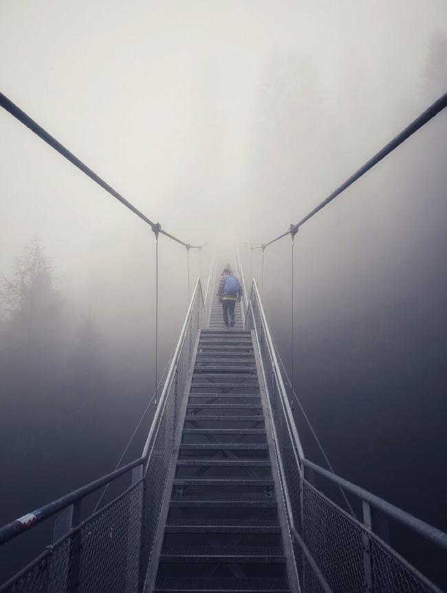 Eine sehr lange wie eine Hängebrücke hängende aufgehängte Metalltreppe führt nach oben in den Nebel wo sie sich verliert. Im Nebel erkennt man links und rechts oberhalb schemenhaft Bäume. Auf der Treppe ist eine Person mit blaumen Rucksack zu erkennen.