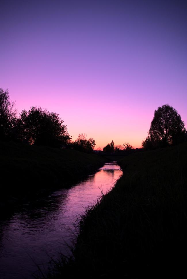 Sonnenuntergang in Blau Lila tönen. Der Himmel spiegelt sich in einem Fluss und alles drum rum ist nur als schwarze Konturen zu erkennen.