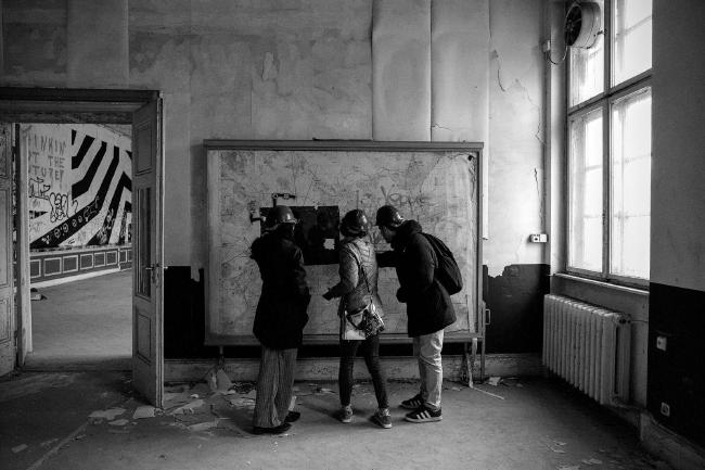 Schwarz-Weiß Bild im Landscape Format. Drei Personen mit Bauhelm stehen vor einer zum Teil beschmierten großen Karte und schauen sich irgendwas darauf an. Der Raum in dem sie sich befinden sieht verwahrlost aus, die Tapeten fehlen zum Teil zum anderen Teil lösen sie sich gerade von der Wand. Von Rechts scheint weiches Licht durch ein Fenster auf die Szene in der Bildmitte. Links ist eine offene Tür die in einen weiteren großen Raum führt dessen sichtbare Wand ein Streifenmuster ziert.