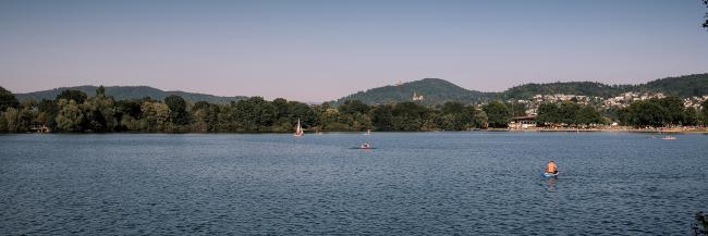 1:3 Farb-Panorama über eine Seefläche mit mehren Menschen auf Standup Boards und auf einem kleinen Segelboot. Die andere Seite des Sees ist im Bäumen bewachsen, wobei im rechen Drittel en Strandbad mit vielen Menschen zu sehen ist. Im Hintergrund sind bewaldete Berge mit Häusern und zwei Burgen