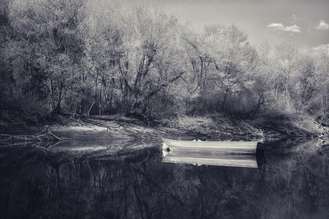Schwarz-Weiß Bild von einem kleinen Boot das auf einer spiegelnden Wasseroberfläche leicht rechts der Mitte schwimmt. Dahinter sieht man von der Sonne hell beschienene Bäume und ein paar kleine Wolken am Stück Himmel den man rechts oben sieht.