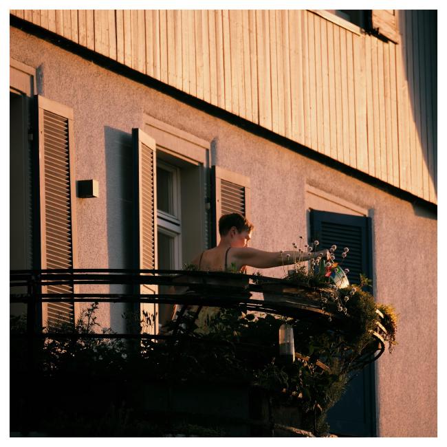 Quadratisches Farbbild. Eine Person mit Trägertopp und kurzen Haaren steht auf einem Balkon und gießt ihre Blumen und ist dabei von der Kamera abgewandt. Dahinter sieht man die Hausfassade die im unteren Teil aus drei Fenstern mit Holzläden und normalem Rauputz besteht und darüber aus Holz. Die Wand leuchtet dabei im Licht der untergehenden Sonne. 