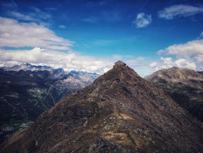 Blick über den Grat zum Brunnenkogelhaus, das auf dessen Spitze liegt. Man sieht im geröll ganz leicht den Weg dort hin. Im Hintergrund liegen Berge auf den anderen Seiten des Tals. Der himmel ist am Rand leicht bewölkt aber sonst blau.