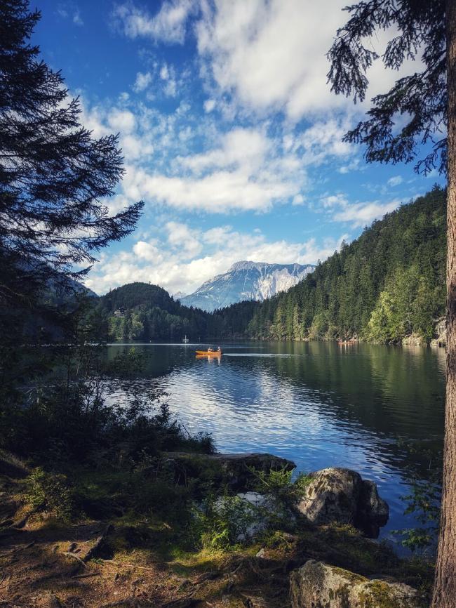 Ein von Bäumen gerahmter See mit einem Holzruderboot in der Mitte. Im Hintergrund verge und Wald. Der Himmel ist blau mit einigen Wolken